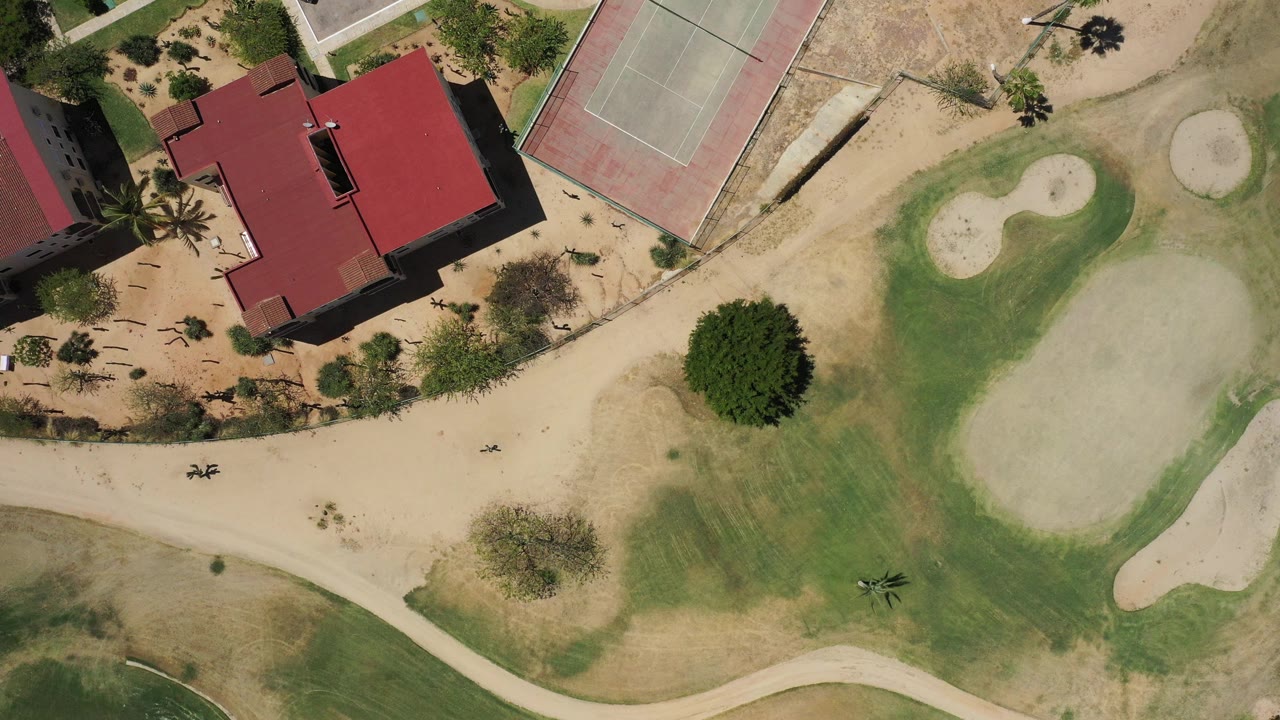 Top-down aerial view of green grounds, sand areas, red-roofed building, and tennis courts.