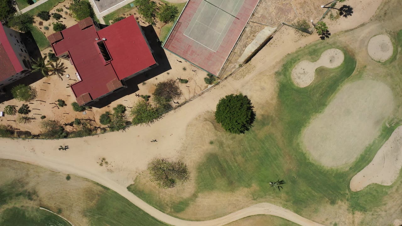 One second into the San Jose del Cabo aerial, the red-roofed structure sits near the upper left while tennis courts, sandy areas, trees, curved paths, and open green grounds fill the frame with no visible people.
