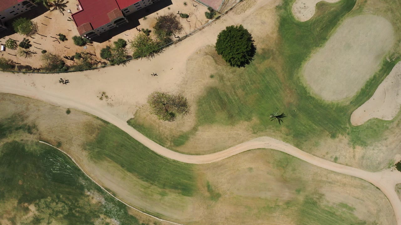 Overhead view in San Jose del Cabo, Mexico showing a curved pale cart path across green and tan golf course turf, with sand bunkers, palms, red-roofed buildings at the top edge, and no visible people.