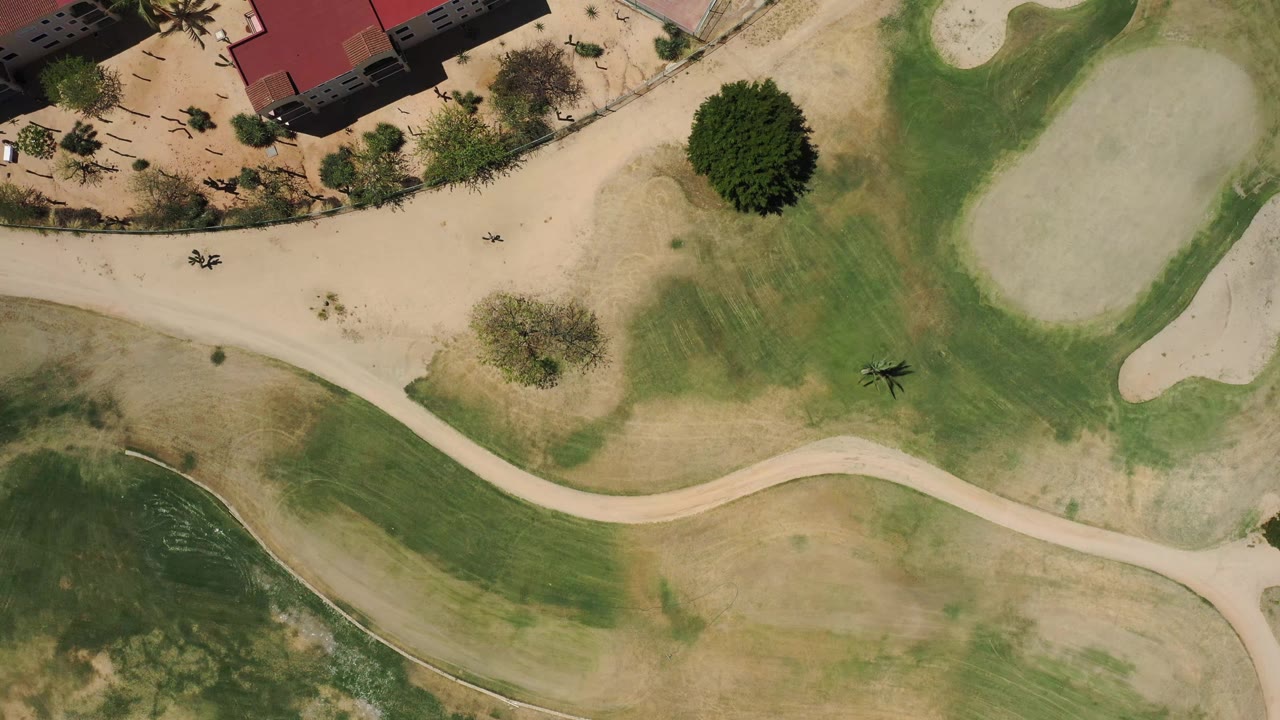 Top-down frame from San Jose del Cabo, Mexico with the fairway and cart paths shifted slightly under the camera, sand traps to the right, palms near the buildings, and no visible people.