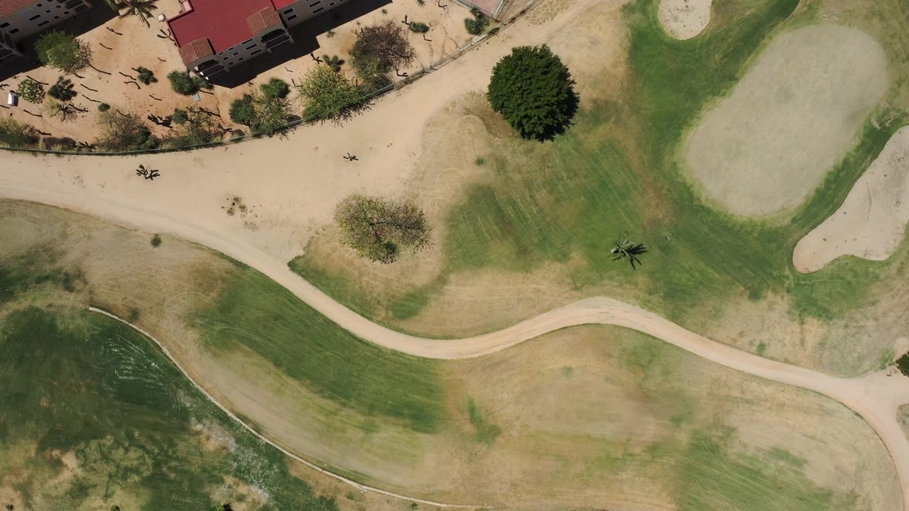 Aerial golf course composition with curved path, green patches, sand bunkers, trees, and red-roofed buildings.