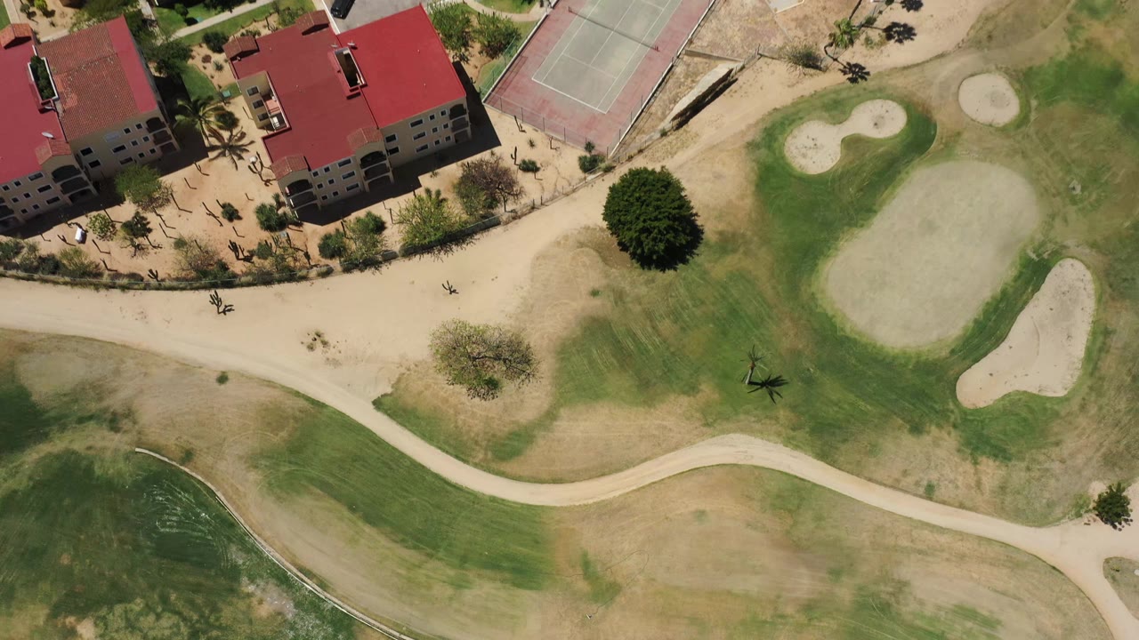 A high overhead view in San Jose Del Cabo shows red-roof buildings at the top of frame, pale cart paths across green and tan turf, sand traps, trees, and no visible people.