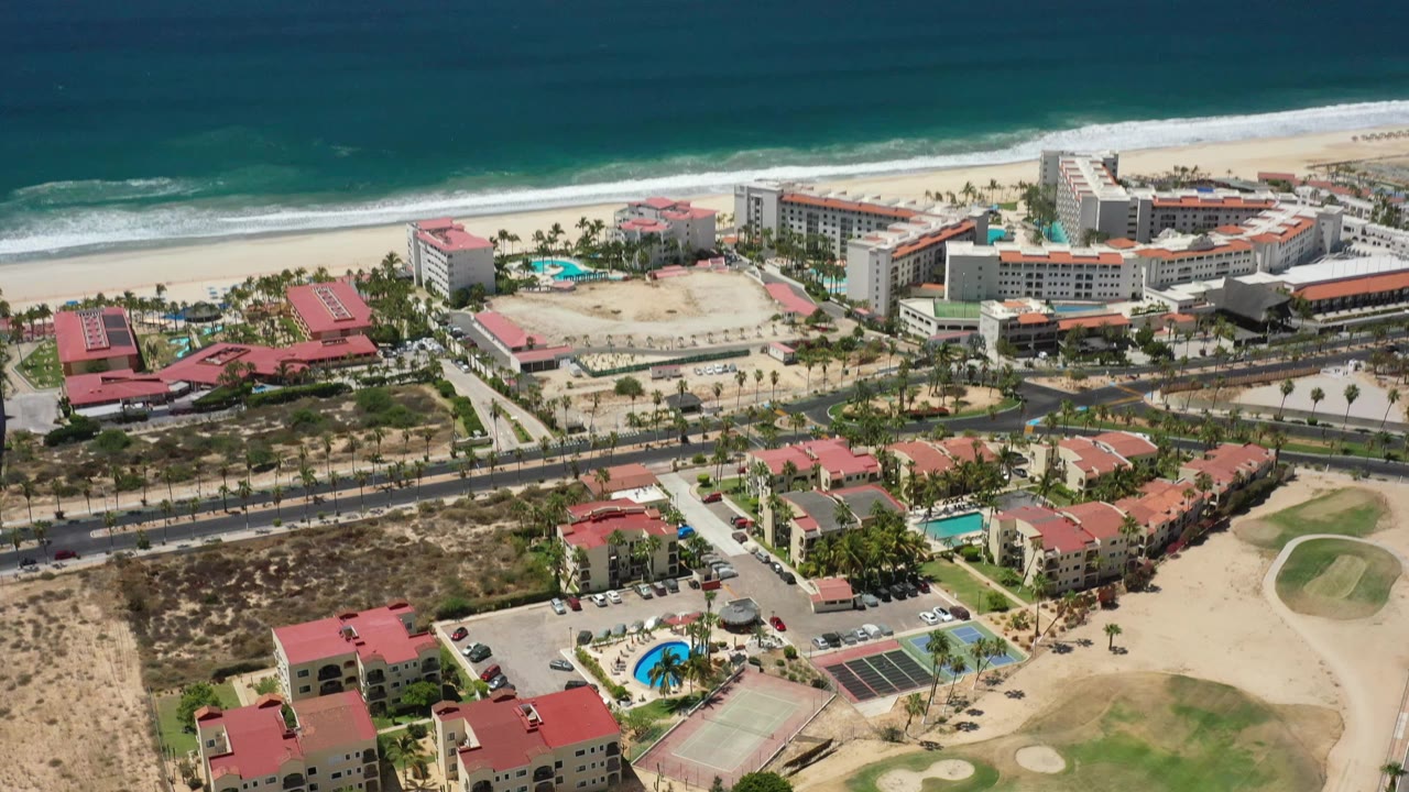 Drone-style aerial frame in San Jose del Cabo showing beachfront hotel blocks, red-roofed villas, pools, palms, roadways, and ocean surf, with no visible people.