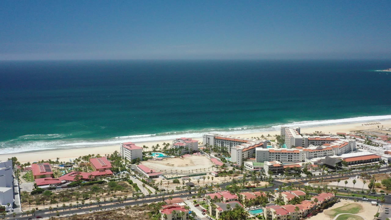 Opening aerial frame over San Jose Del Cabo, Mexico, showing blue-green ocean, a white surf line, beachfront hotels, roads, palms, and red-roofed buildings with no visible people.