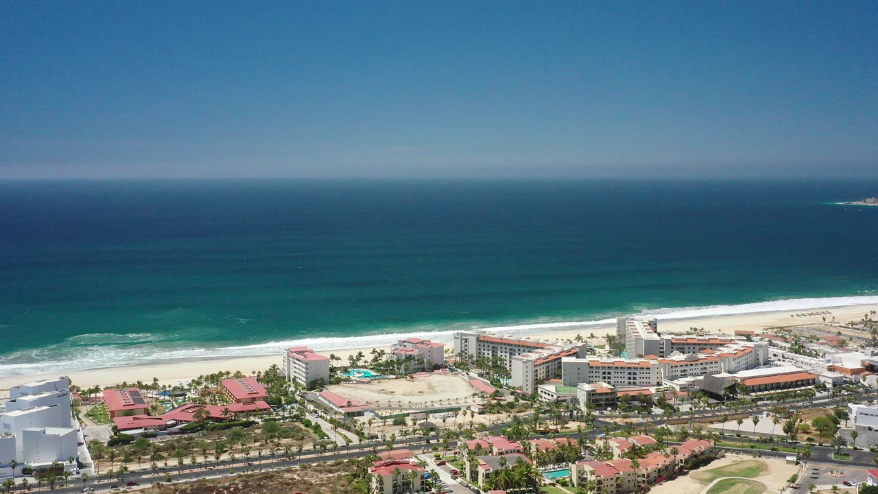 High aerial view over San Jose del Cabo with blue ocean, white surf, sandy beach, resort buildings, palms, and no visible people at the start of the clip.