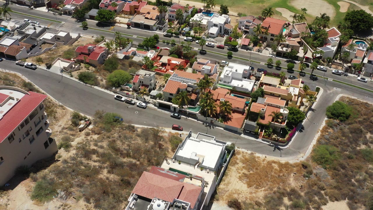 High oblique aerial view of San Jose del Cabo residential streets, modern homes, parked vehicles, dry scrub lots, and green landscaping at the top edge, with no visible people.