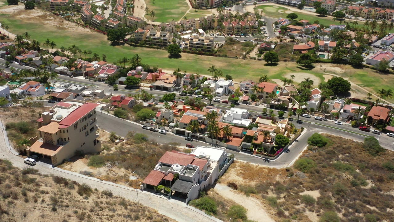 Oblique aerial view in San Jose del Cabo, Mexico, with dry scrub lots in the foreground, homes and roads in the midground, green grounds beyond, and no visible people.