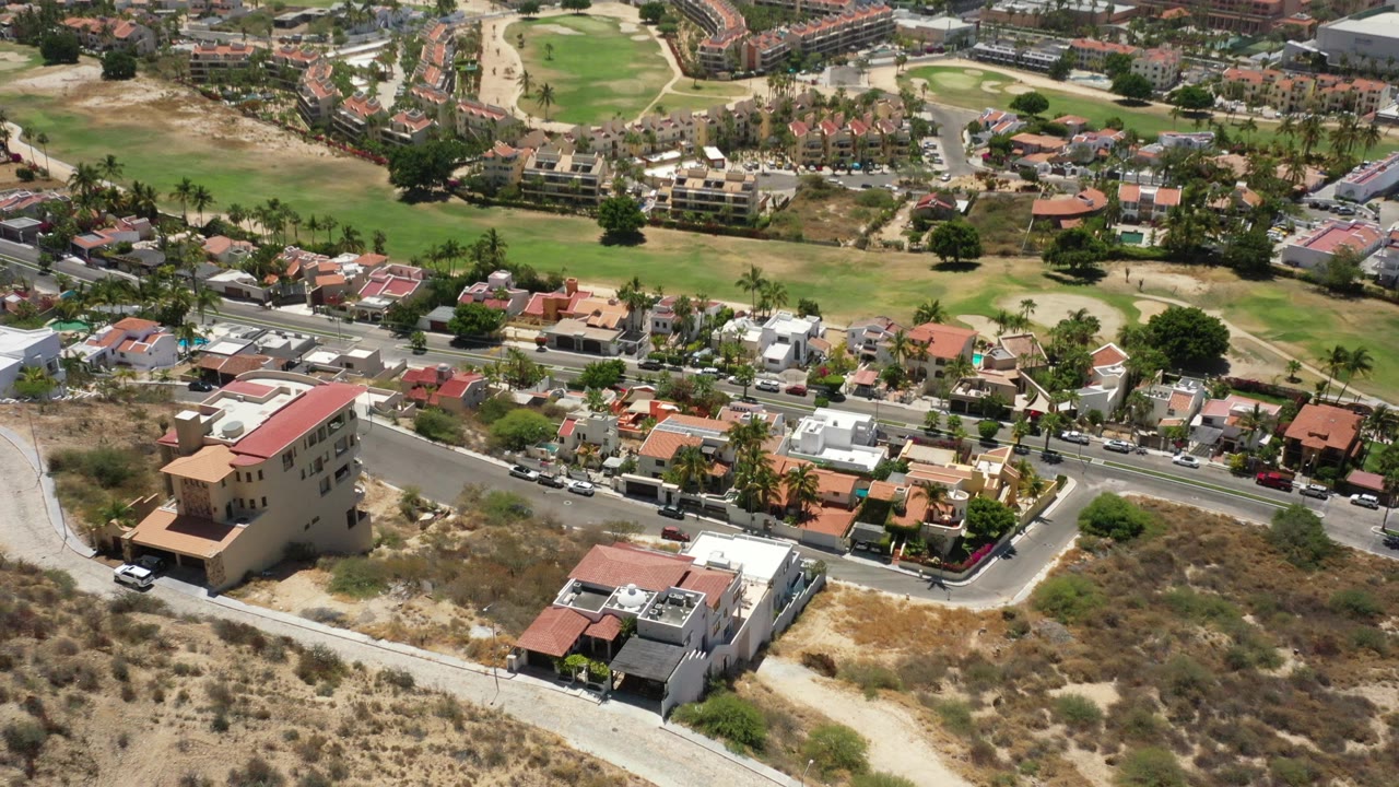 High aerial view of homes, roads, green grounds, and coastal buildings in bright sun.