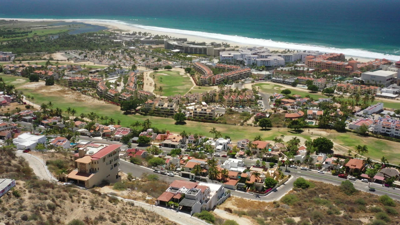 Aerial view over San Jose del Cabo showing dry foreground land, rows of houses, larger beachfront buildings, pale sand, blue ocean, and no visible people.