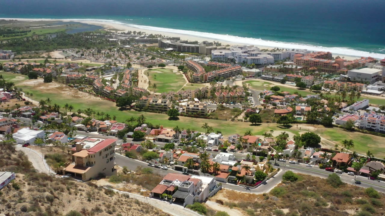 Wide San Jose del Cabo aerial frame with homes and roads in the foreground, green landscaped grounds in the center, beachfront buildings and ocean beyond, and no visible people.