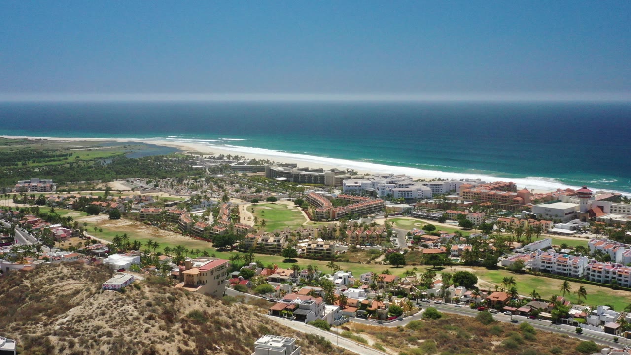 High aerial view over San Jose Del Cabo with dry hillside foreground, resort buildings, golf greens, sandy beach, blue ocean, and no visible people.