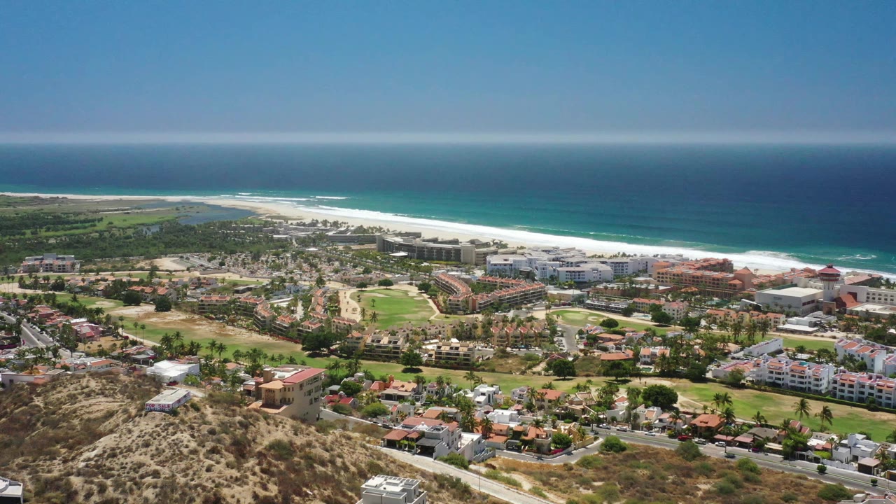 Aerial frame of San Jose Del Cabo showing terracotta roofs, white resort blocks, landscaped fairways, a long beach, breaking surf, and no visible people.