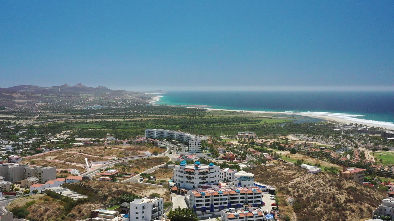 Wide aerial view over San Jose Del Cabo with white hillside buildings in the foreground, green land beyond, a long pale beach, blue ocean, and no visible people.