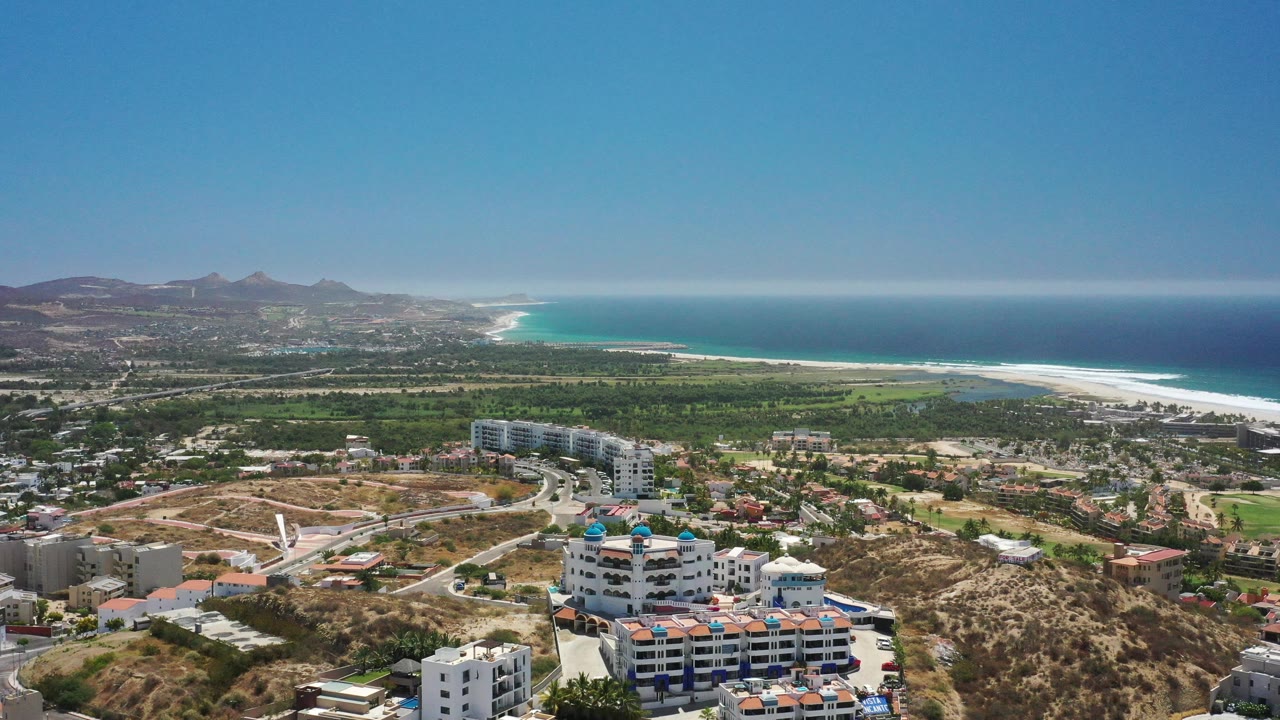 Aerial frame from San Jose Del Cabo showing apartment blocks and winding roads below, green terrain across the middle distance, white surf along the beach, and no visible people.