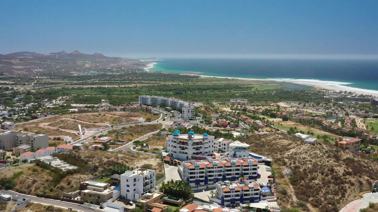 Aerial view in San Jose del Cabo showing white resort blocks below, a curving road through dry terrain, green flats near the beach, and no visible people.