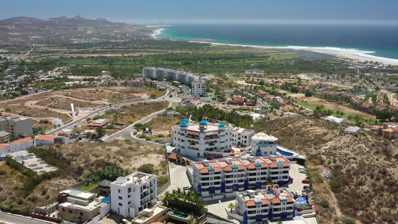Aerial view over San Jose Del Cabo resort buildings with blue domes, dry hillside roads, green lowland, and a bright ocean shoreline in the distance, with no visible people discernible.