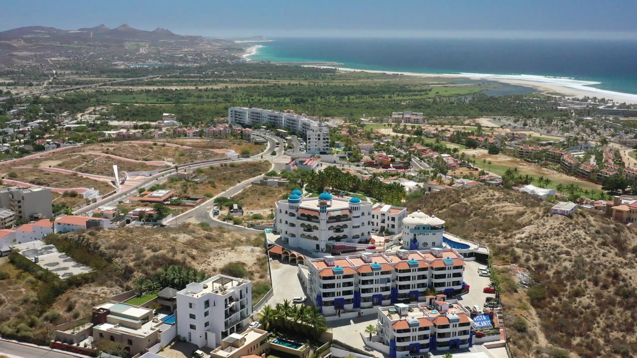 High aerial frame in San Jose Del Cabo showing white resort blocks at lower center, curved roads across dry terrain, green vegetation beyond, and pale surf along the distant coast, with no visible people discernible.