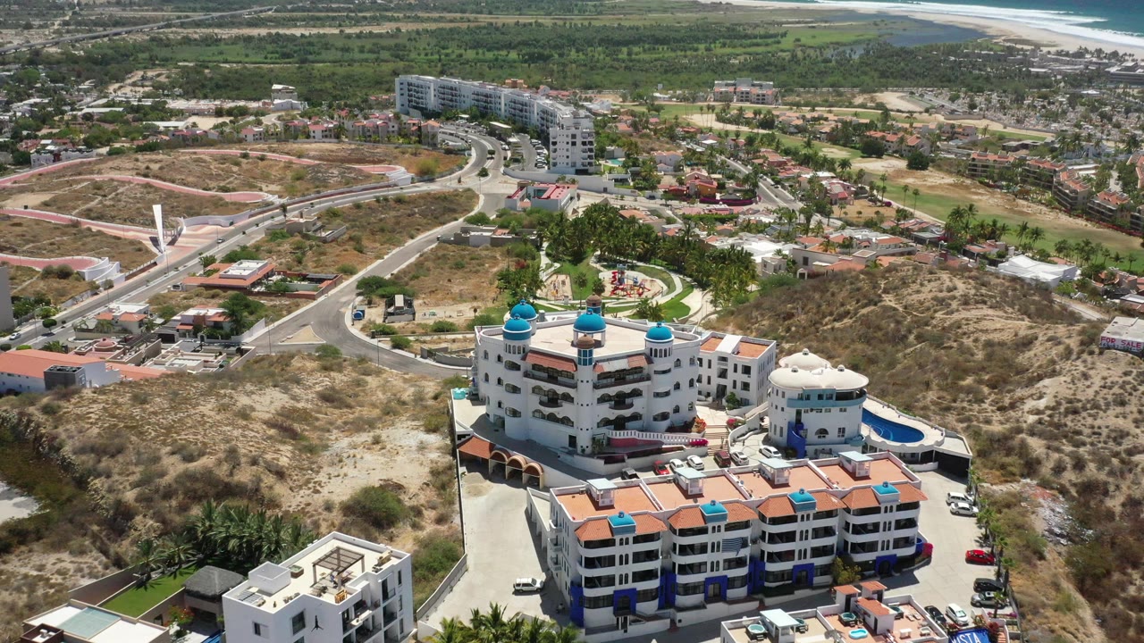 A slightly shifted aerial frame over San Jose del Cabo shows the blue-domed building lower in view, a looping road system, scattered homes, scrub-covered hills, distant beach, ocean, and no visible people.