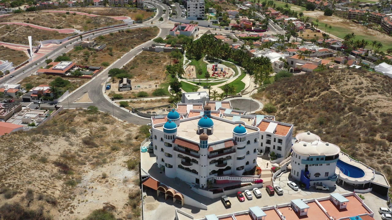 Aerial frame over a white resort with turquoise domes in San Jose del Cabo, Mexico, with dry hillside, roads, scattered homes, and no visible people.