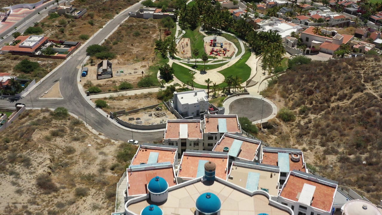Aerial frame over San Jose del Cabo showing flat-roofed homes at the bottom, a curved road, dry sandy land, and a small green park with paths and playground equipment in bright sun.