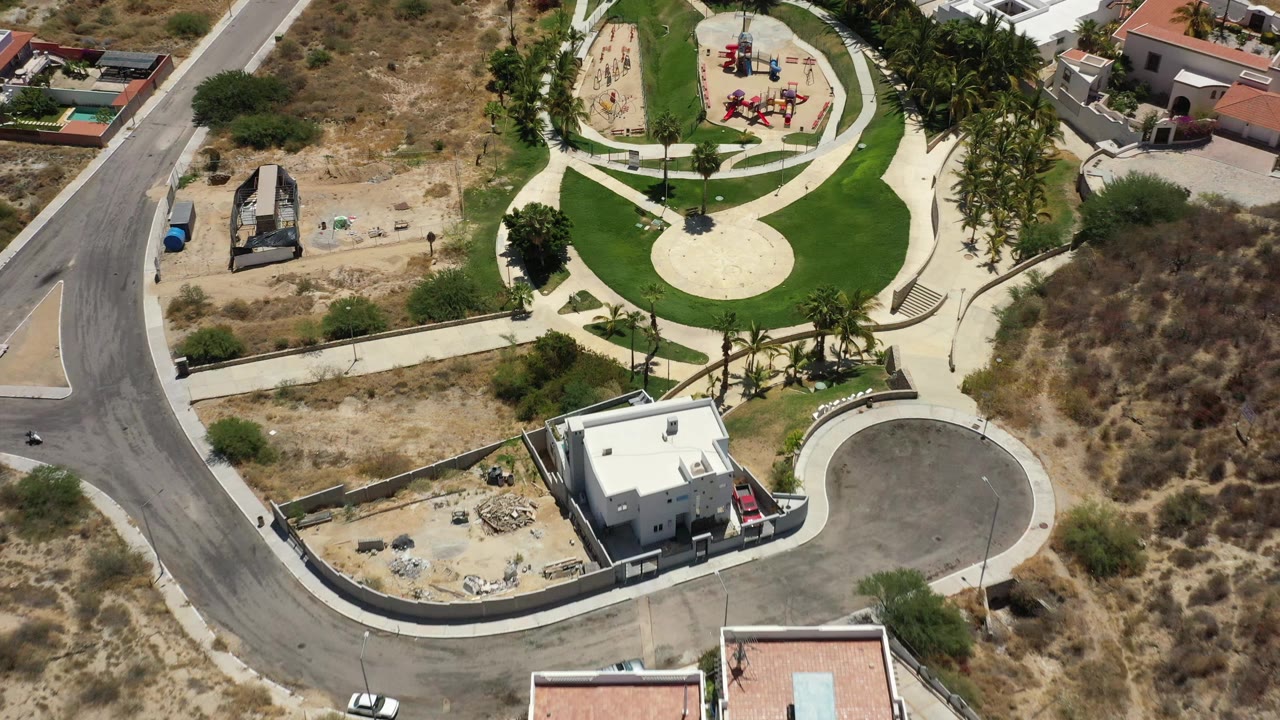 Aerial frame of the San Jose Del Cabo park with the circular pale play surface, red playground structures near the top, palm trees, streets, a white-roofed house, and tiny visible people.