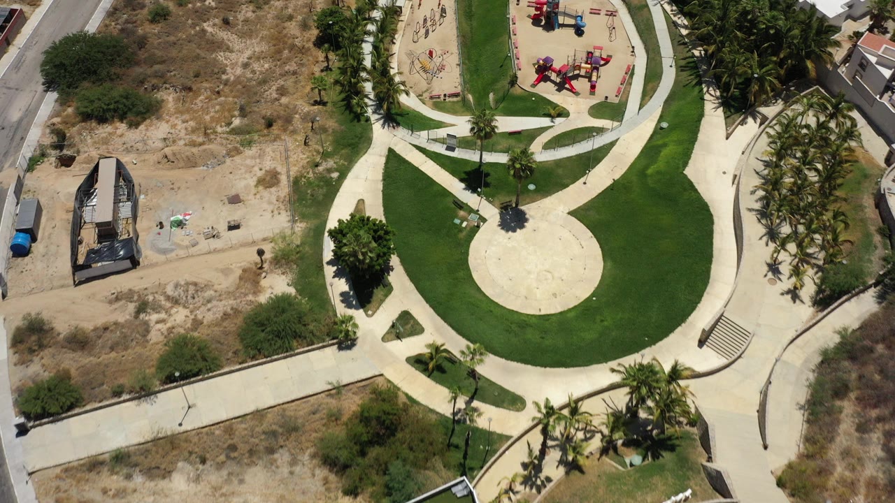 Overhead view of a San Jose del Cabo park at 0:00, showing a circular paved area, green lawn, pale walkways, playground equipment, palm trees, dry scrubland, and tiny visible people.