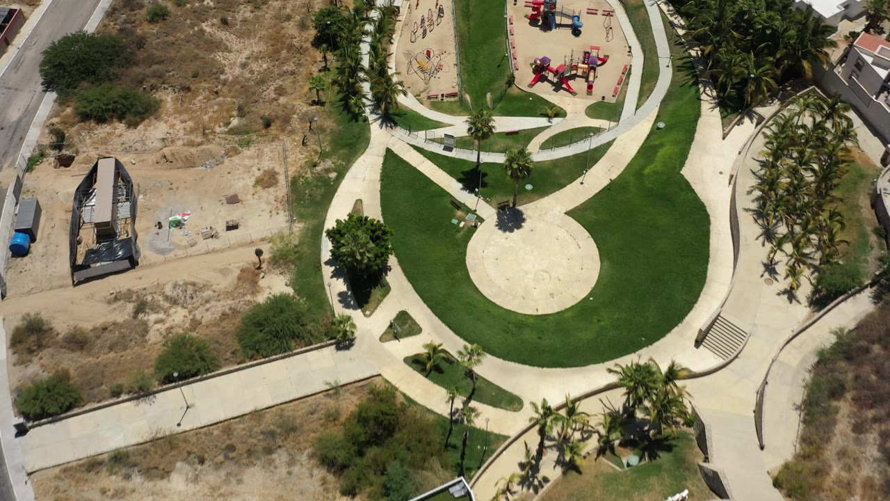 Aerial frame from San Jose del Cabo at 0:01 with the circular plaza centered low, the playground near the upper right, palms along the right edge, dry ground to the left, and tiny visible people.