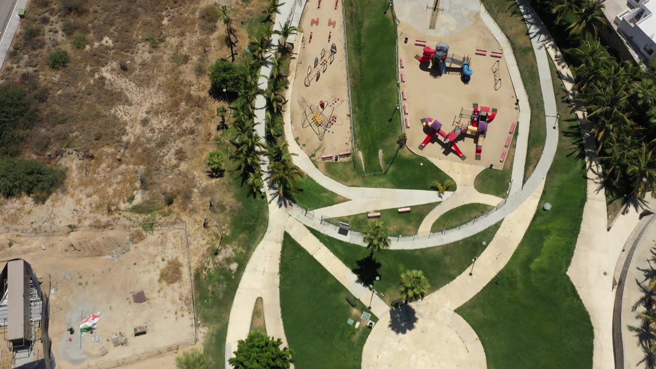 Top-down view of a landscaped park with lawns, paths, palms, dry ground, and playground equipment.