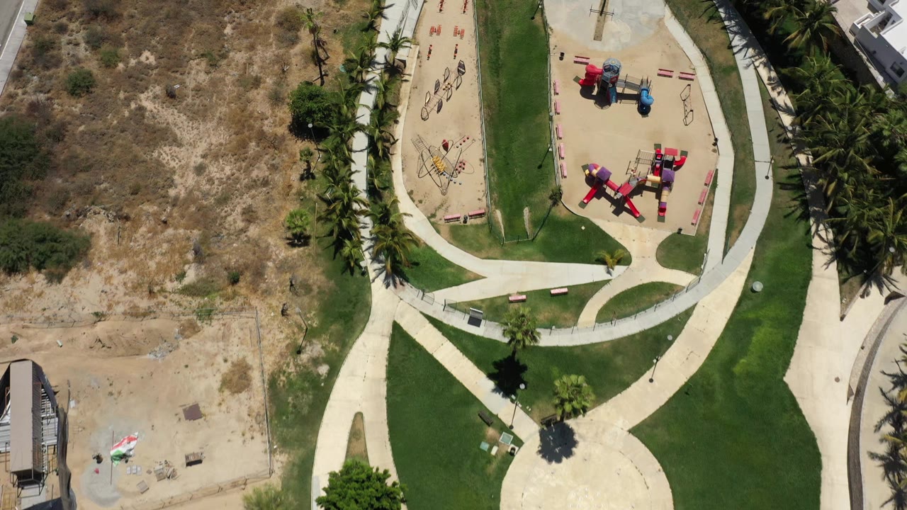 Second overhead frame in San Jose del Cabo, Mexico with the playground shifted slightly right in frame, bordered by palms, mowed lawn, dry open ground, and pale walkways.