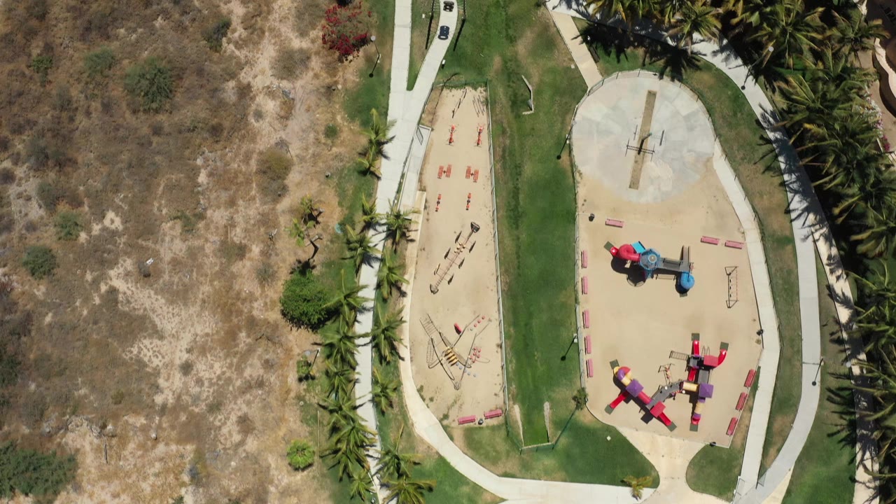 Aerial view in San Jose del Cabo centered on red and blue playground equipment, tan ground cover, a lawn strip, pale walkways, and dense palms along the right side, with no visible people.