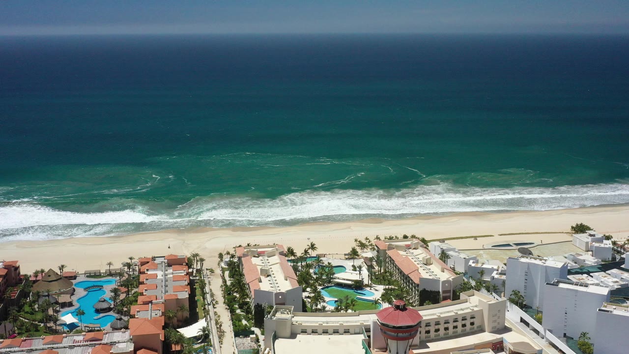 Aerial frame over San Jose del Cabo showing dark blue ocean, turquoise nearshore water, white surf, pale sand, resort buildings, pools, palms, and no visible people.