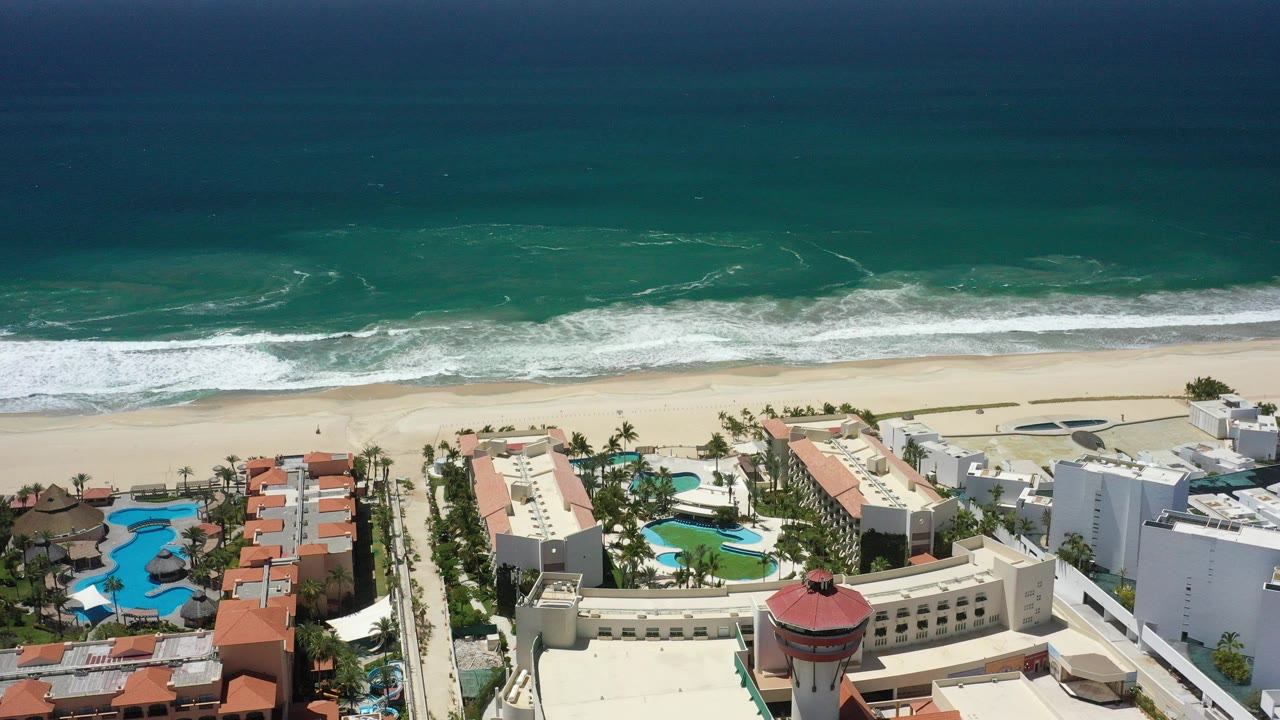 Opening aerial frame over San Jose del Cabo resort rooftops, pools, palm rows, sandy beach, and turquoise ocean surf with tiny beachgoers visible.
