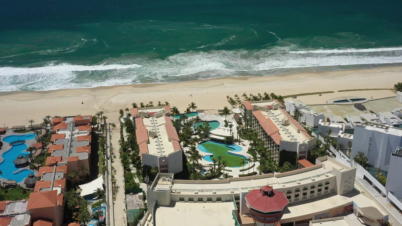 Aerial view over a San Jose del Cabo beachfront resort with red tile roofs, curving blue pools, a sandy beach, rolling surf, and no visible people.