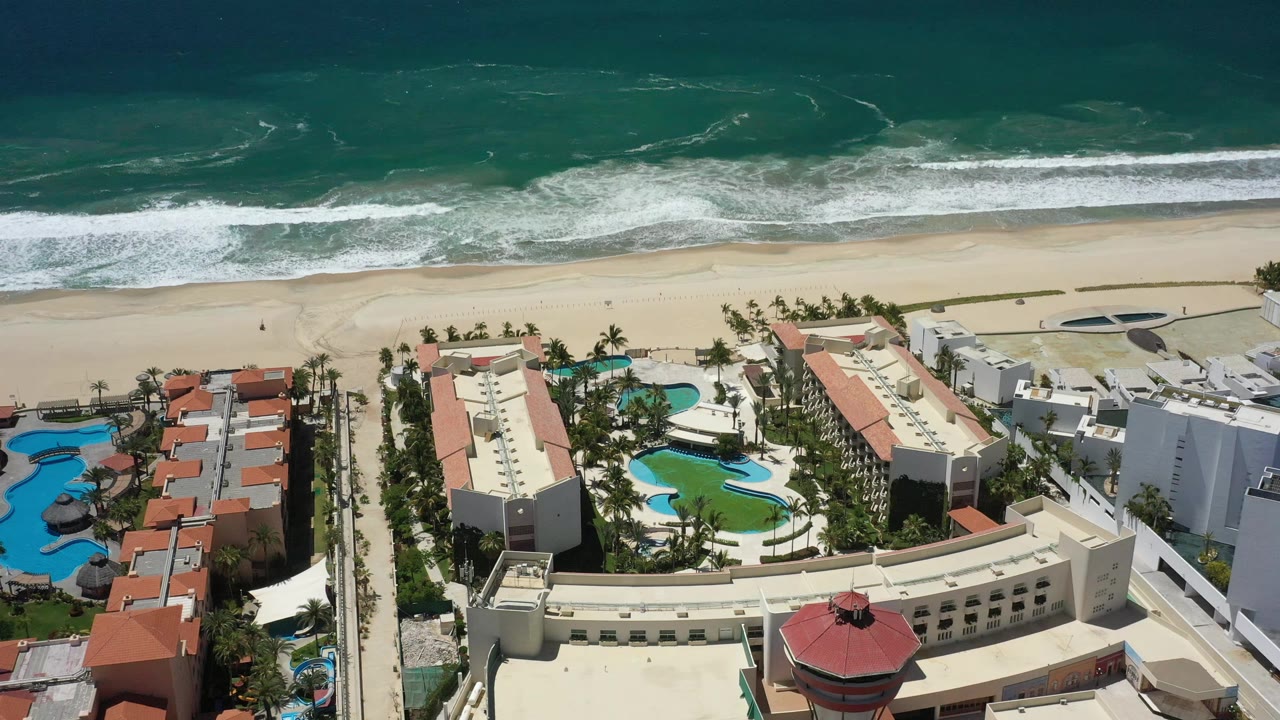 High aerial frame in San Jose del Cabo showing the beach line higher in view, white surf bands offshore, resort roofs, pools, palms, and no visible people.
