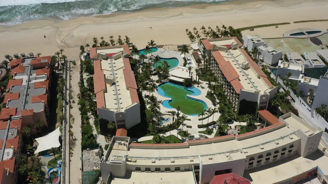 A high aerial view in San Jose Del Cabo shows resort buildings around curved swimming pools, palm trees, sandy beach, and surf at the top of frame, with no visible people.