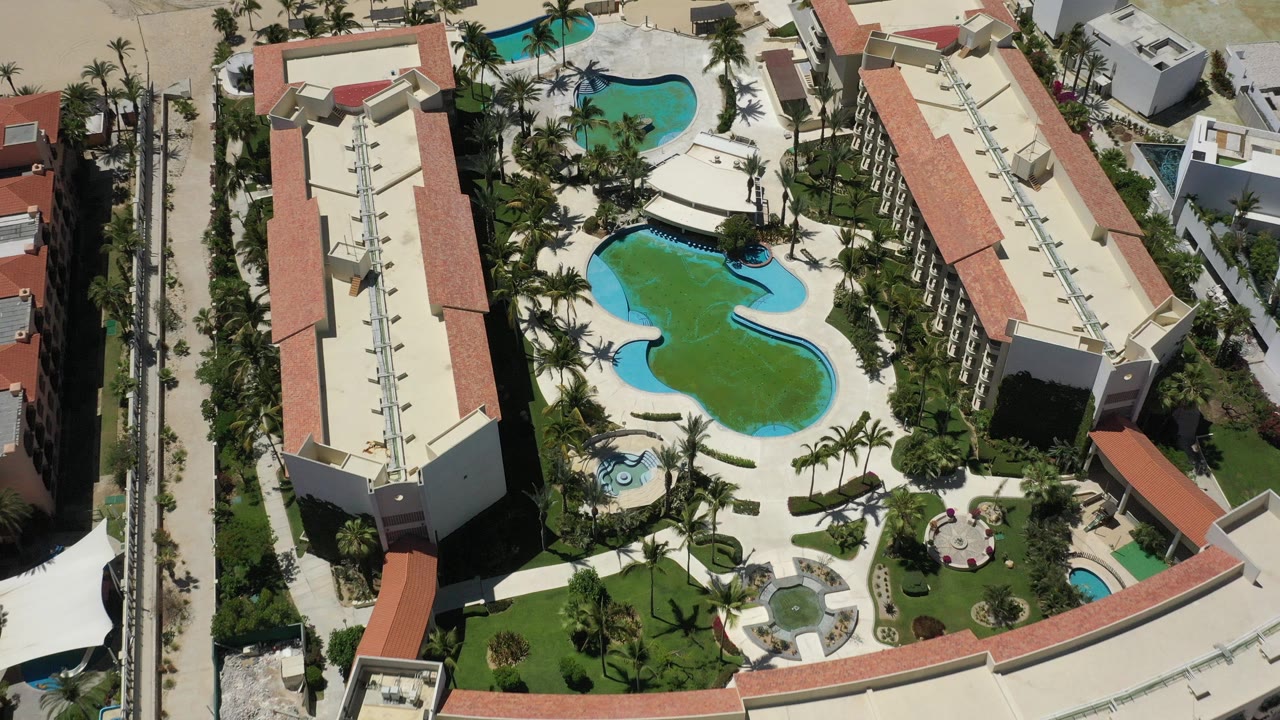 Aerial view over a San Jose del Cabo resort with two long red-roof buildings, curved blue pools, palm landscaping, and no visible people.