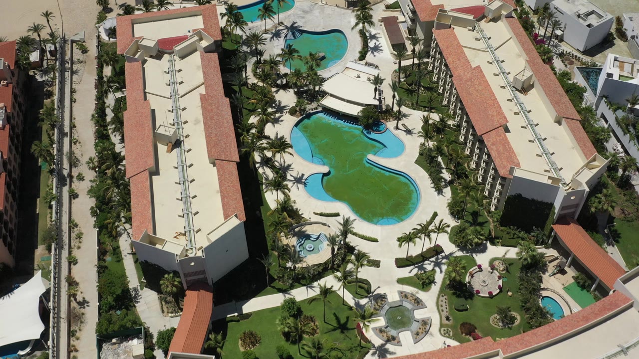 Top-down view of resort pools, palms, and hotel roofs near beach sand.