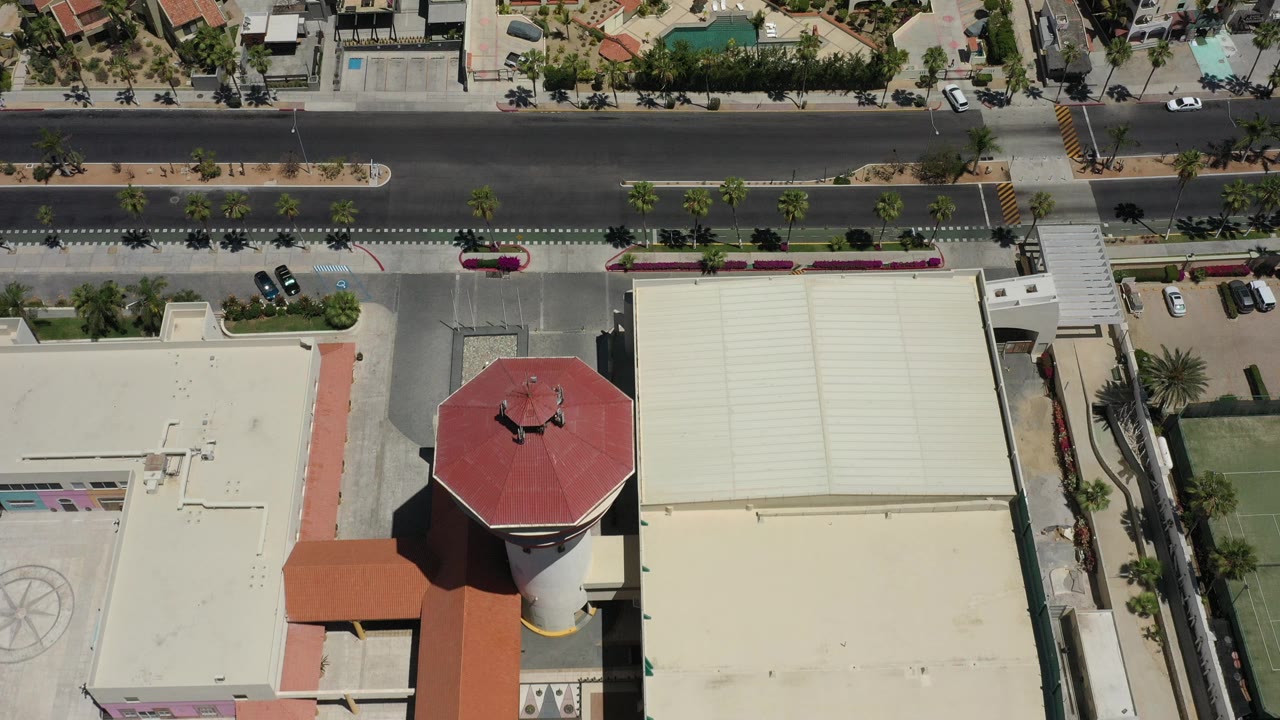 High overhead frame in San Jose del Cabo showing a wide road, red-tile and flat rooftops, palm-lined sidewalks, turquoise pools, parked cars, and no visible people.