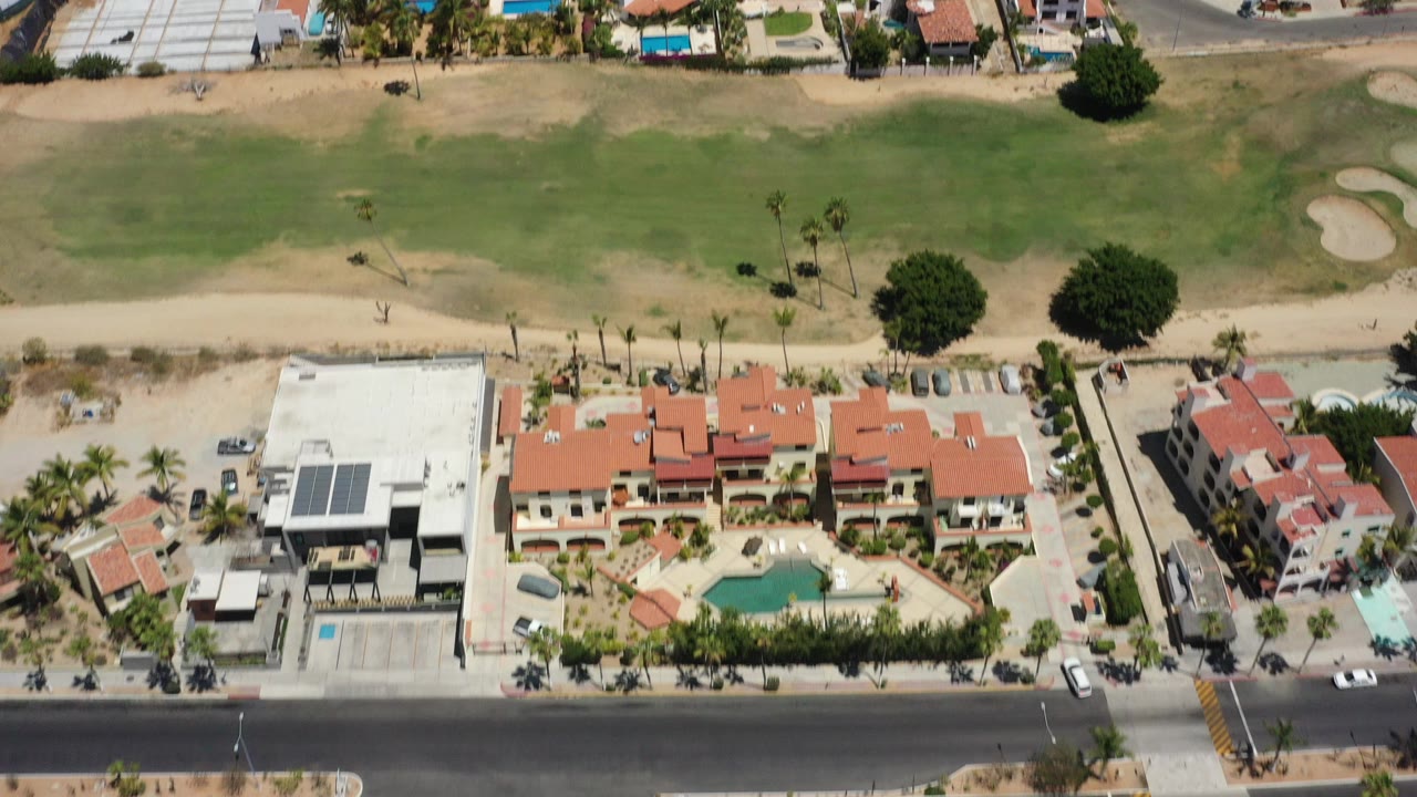 High overhead view in San Jose Del Cabo showing red-roof homes, a roadside row of buildings, pools, palm trees, and green course-like land with no visible people.