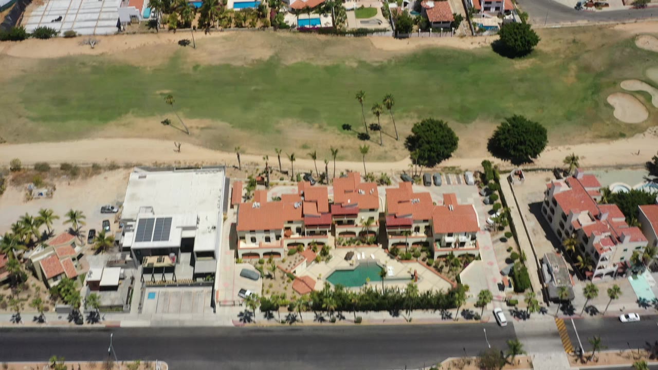 Overhead San Jose Del Cabo frame with a broad green field at top, sandy path, red-roof homes, a white building with solar panels, a road, and no visible people.