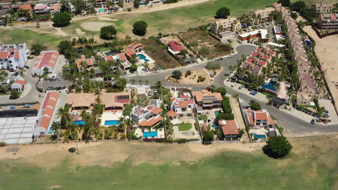 Aerial frame over San Jose del Cabo showing red-roofed homes, blue pools, palm trees, a paved road, and green open ground at the top, with no visible people.