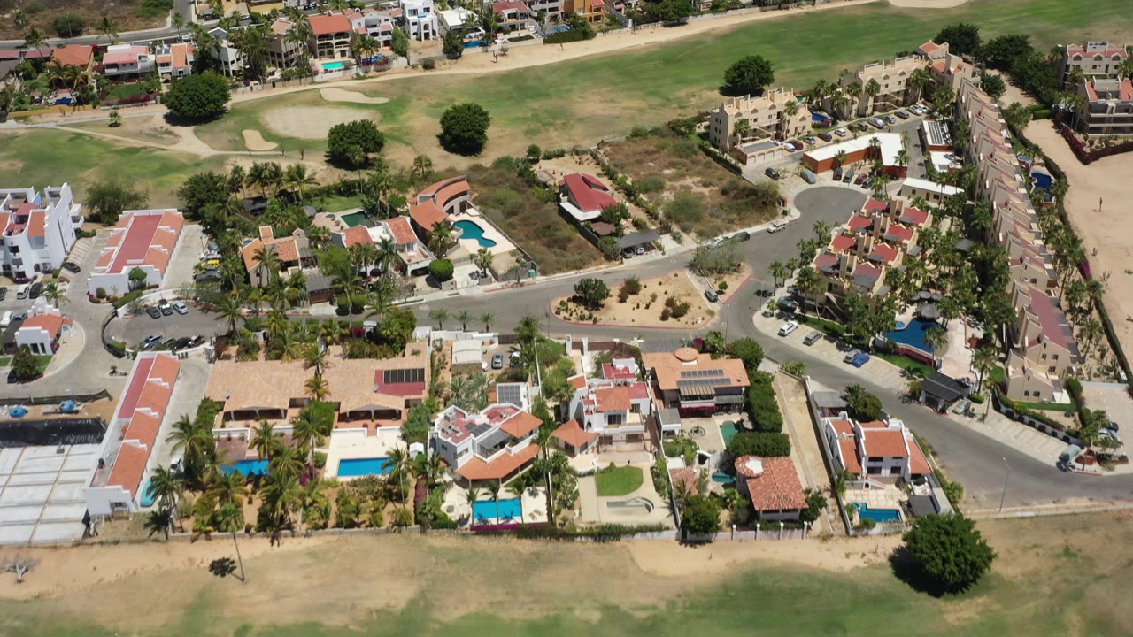 High aerial of residential streets, pools, palms, and green grounds beyond the homes.