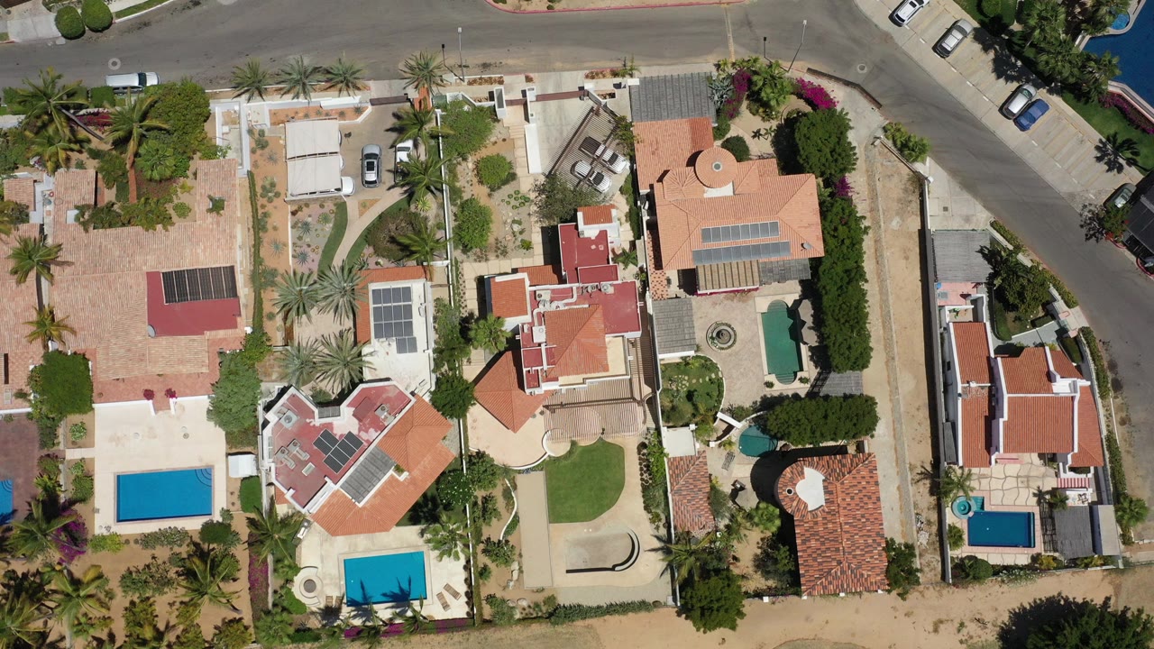 Top-down aerial frame over a San Jose del Cabo residential block with tan roads, red-roofed homes, blue swimming pools, palm trees, parked vehicles, and no visible people.
