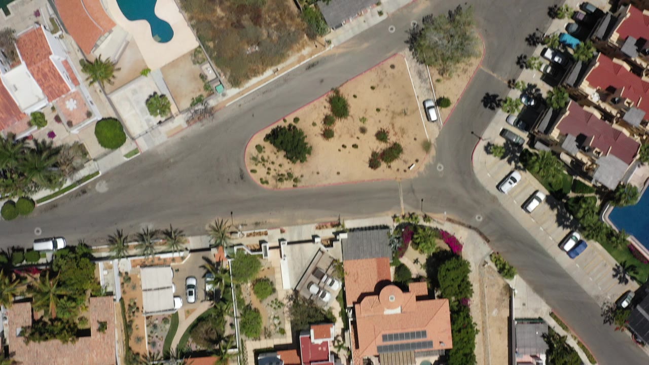 Top-down aerial frame over San Jose del Cabo showing intersecting residential streets, red-roof homes, parked cars, desert yards, palm trees, and a blue swimming pool with no visible people.