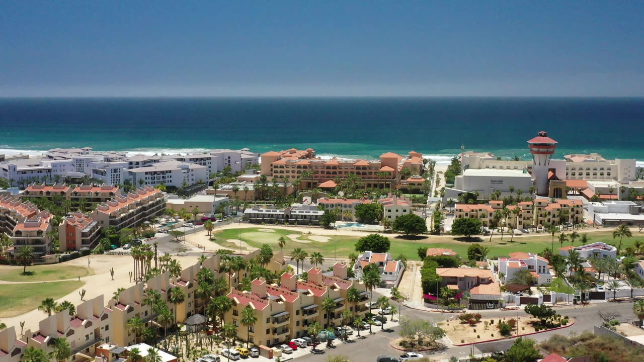 Aerial frame in San Jose del Cabo, Mexico, with the red-topped tower near the right side, low buildings below, landscaped green space, surf line, and no visible people.