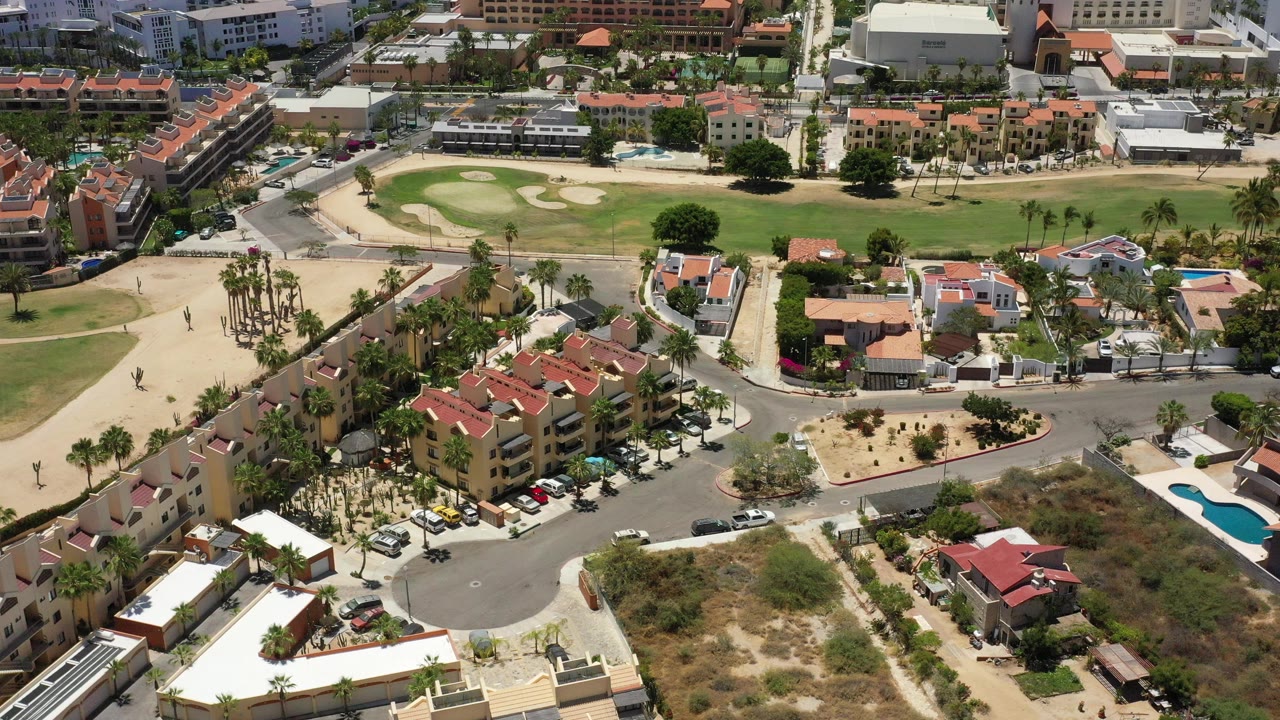 Opening aerial frame over San Jose del Cabo, Mexico, showing red-roof homes, a curved street, parked cars, palm trees, pools, dry lots, and a green course area, with no visible people.