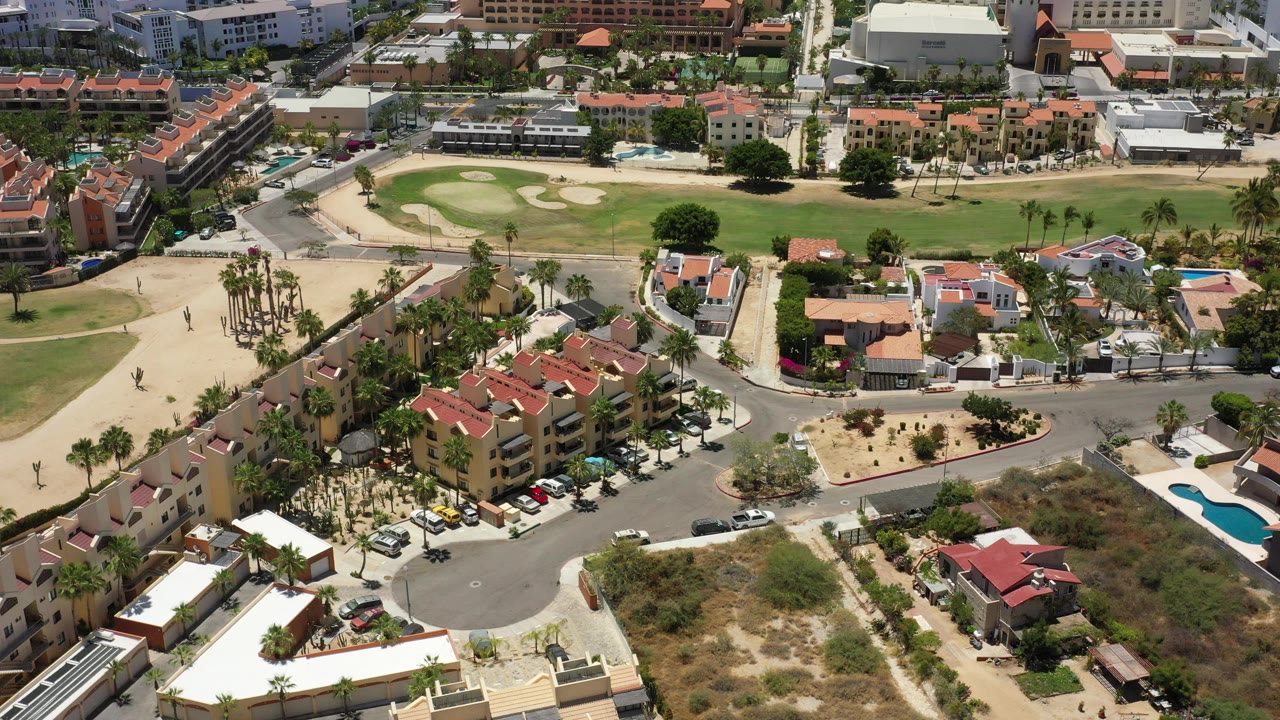 Second frame of the San Jose del Cabo aerial holds over a cul-de-sac, tan and red-roof buildings, palm rows, small pools, parked cars, and the green open area beyond, with no visible people.