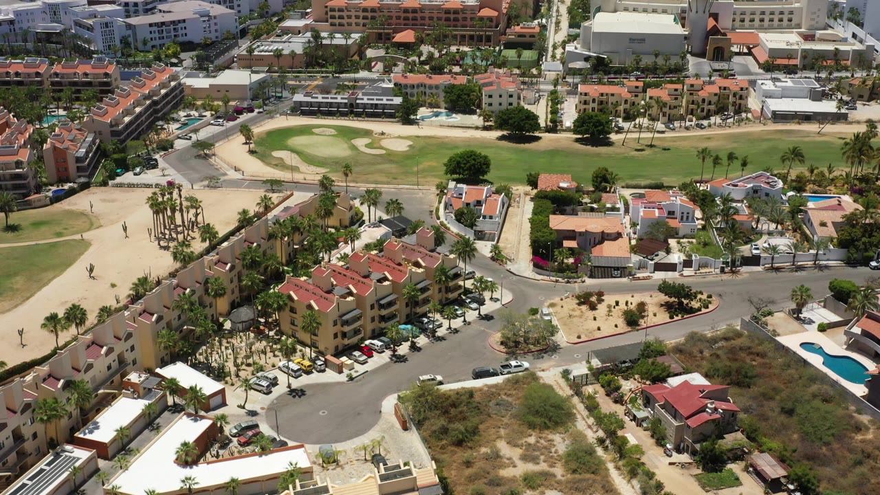 Aerial view in San Jose del Cabo with resort rooftops in the foreground, a broad green space near the center, beach and ocean behind, and no visible people.
