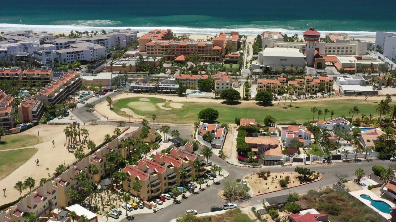 Opening aerial frame over San Jose Del Cabo with blue ocean, white surf, beachfront buildings, a green lawn, red roofs, streets, pools, and no visible people.