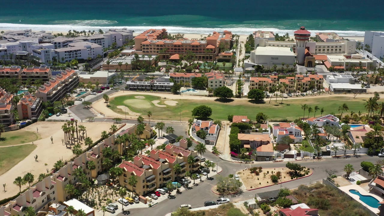 Aerial frame in San Jose Del Cabo showing the ocean band higher in frame, white surf behind hotel buildings, palms, roads, red-roofed structures, and no visible people.